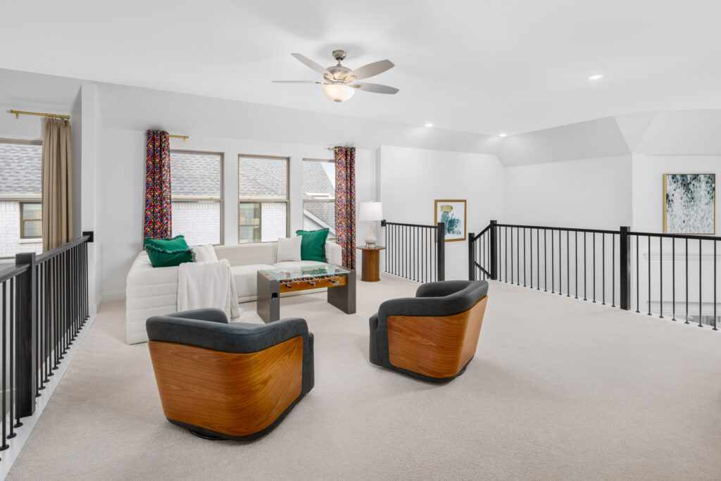 A modern loft living area in Painted Tree with a white sofa, two wood-accented chairs, a coffee table, and large windows with colorful curtains. Black railings line the open space; Coventry Homes brings McKinney style to life here.