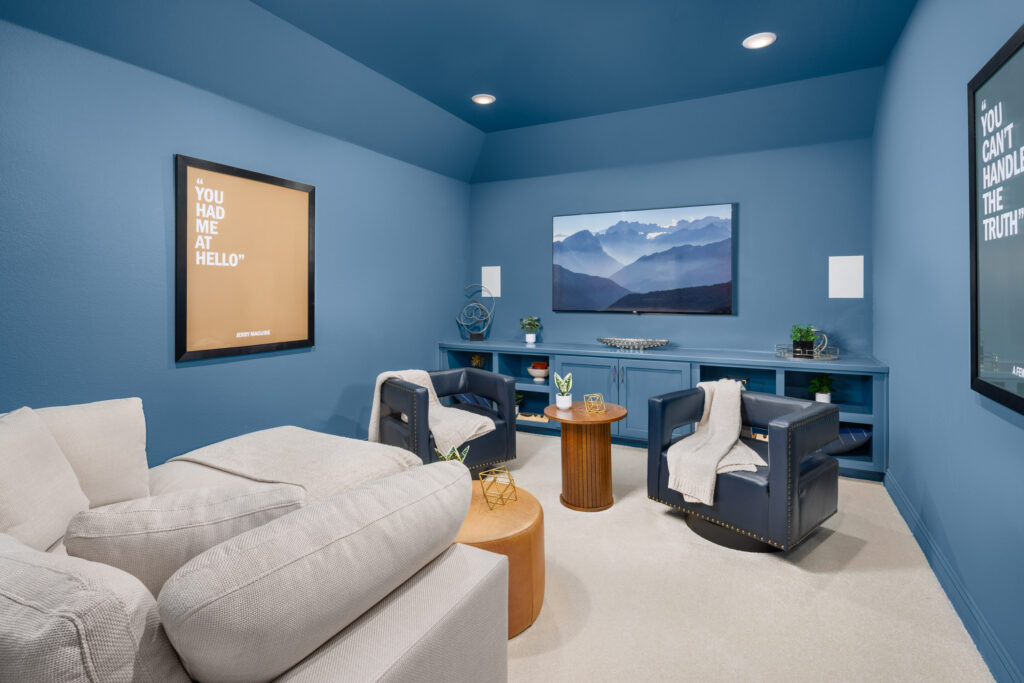 Modern home theater room in a new home at Painted Tree, McKinney, featuring blue walls, a wall-mounted TV, two navy chairs, a beige sectional sofa, framed posters, and a small wooden table.