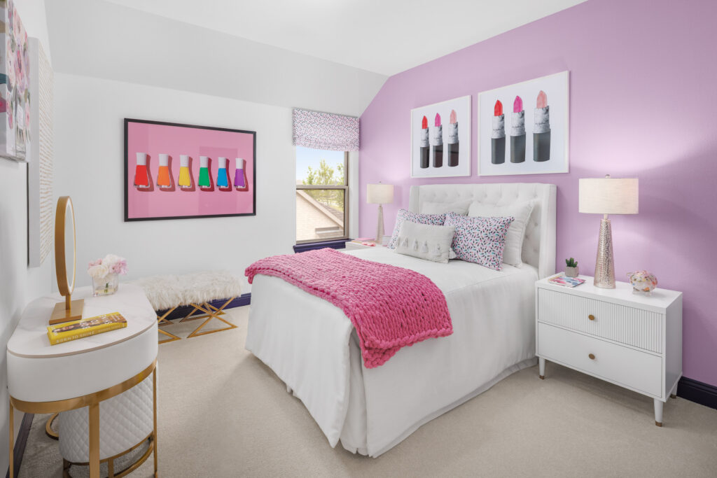A bedroom in a new home with white and lavender walls, a bed with a pink blanket, makeup-themed wall art, white furniture, and a window letting in natural light—perfect for relaxing in Painted Tree by Coventry Homes.