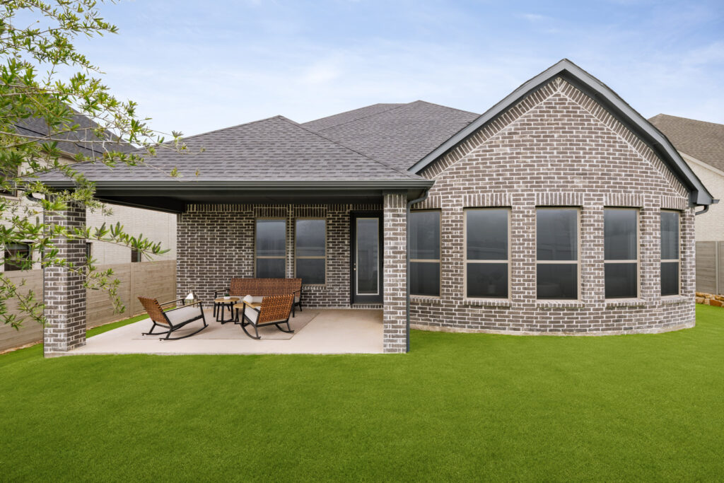 Back view of a modern new home in McKinney, TX, featuring a gray brick exterior, large windows, and a covered patio with outdoor furniture on a green lawn.