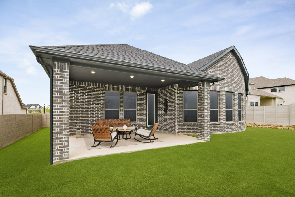 Covered brick patio with outdoor seating and table, attached to a modern McKinney, TX home with large windows, overlooking a well-kept lawn under a partly cloudy sky.