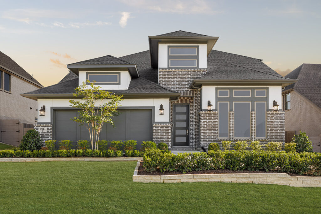 Two-story modern new home in McKinney’s Painted Tree, featuring gray brick and siding exterior, double garage doors, a landscaped front yard, and large windows.