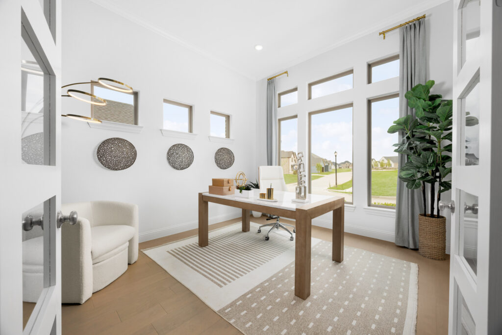 Modern home office in TX’s Painted Tree community, featuring a wooden desk, white chair, computer monitor, large windows, patterned rug, wall art, and a tall potted plant—perfect for your new home workspace.