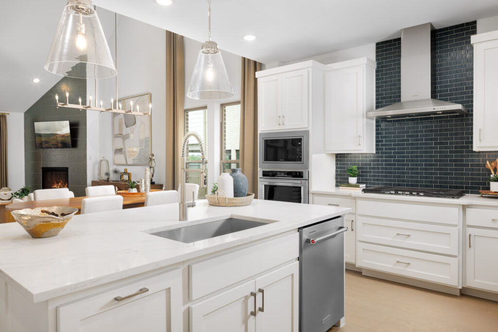 Modern kitchen in this Coventry Homes new home features white cabinets, a spacious island with sink, stainless steel appliances, and dark subway tile backsplash; open dining area visible in the background. Perfect for TX living.