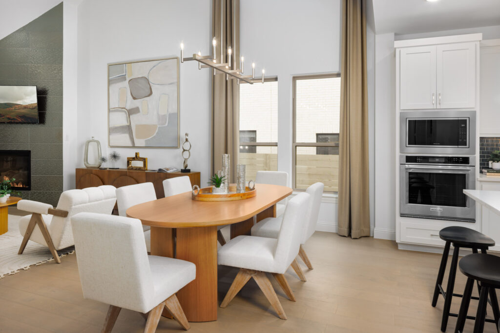 Modern dining area in a McKinney new home at Painted Tree, featuring a wooden oval table, six upholstered chairs, sideboard, abstract art, and chic light fixture, adjacent to a kitchen with built-in appliances and bar stools.