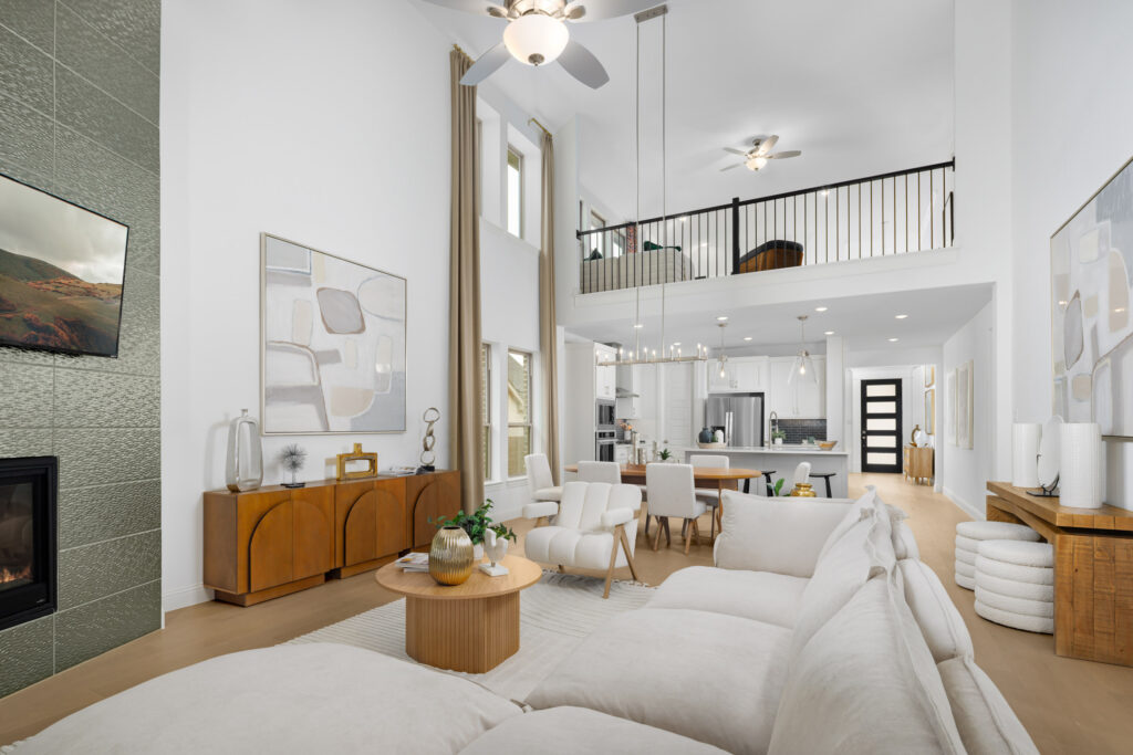 Modern open-concept living room in a new TX home at Painted Tree, featuring a white sectional sofa, dining area, kitchen, large windows, and a loft above. Neutral tones and contemporary decor throughout.
