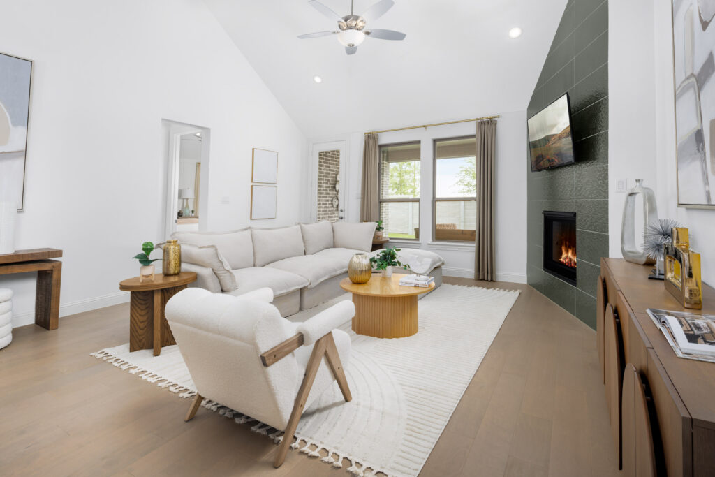 Modern living room in McKinney, TX by Coventry Homes featuring white walls, beige furniture, a round wooden coffee table, fireplace with green tile and TV above, large windows, and light wood flooring.