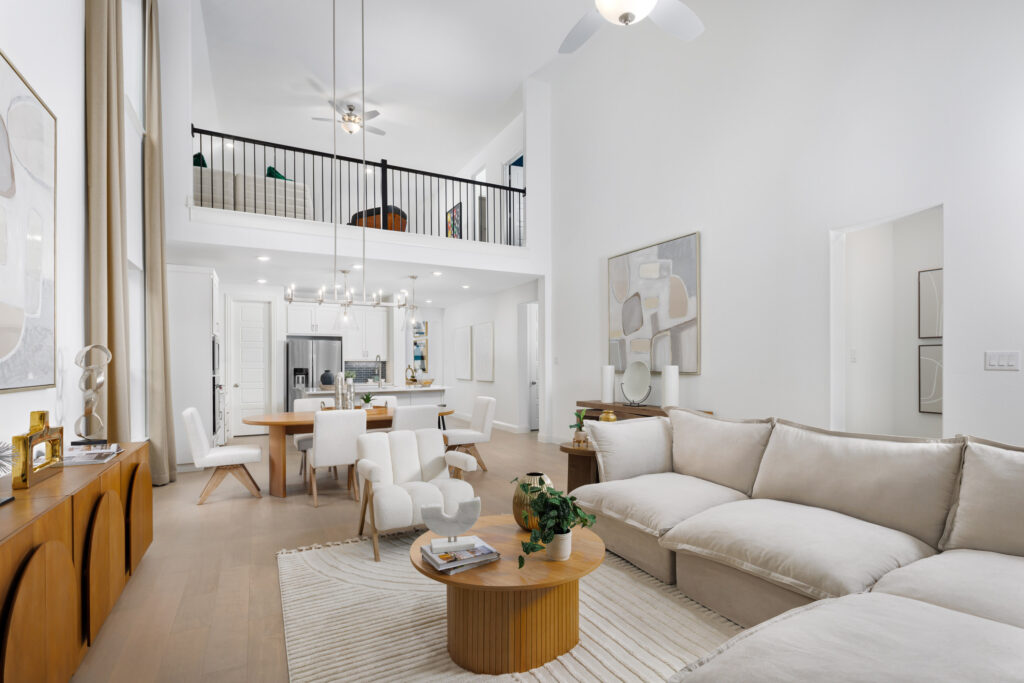 Modern open-concept living area in a new home at Painted Tree, featuring neutral decor, sectional sofa, dining table, kitchen, and loft with sleek black railing above.