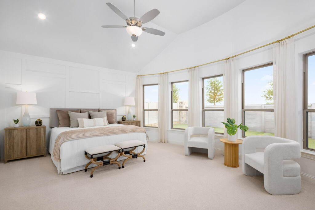 Bright, modern bedroom in a new Coventry Homes TX home features a large bed, two nightstands, two white chairs, a small round table, large windows, and a ceiling fan.