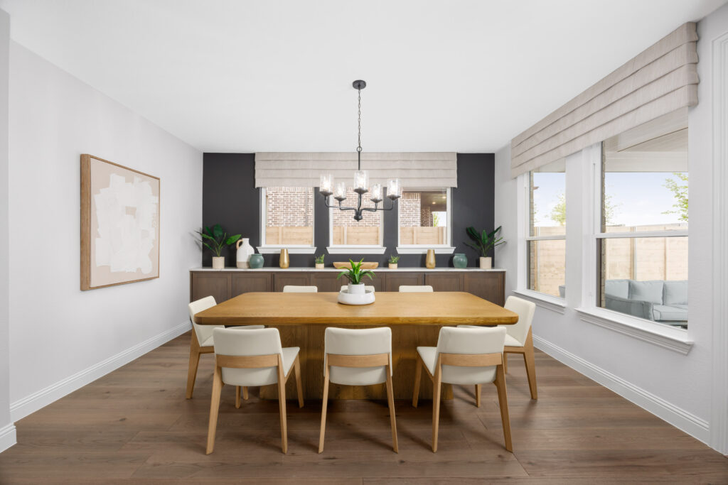 Modern dining room in a new home at Painted Tree, McKinney, featuring a wooden table, eight white chairs, large windows, neutral walls, abstract artwork, and a chandelier hanging from the ceiling.