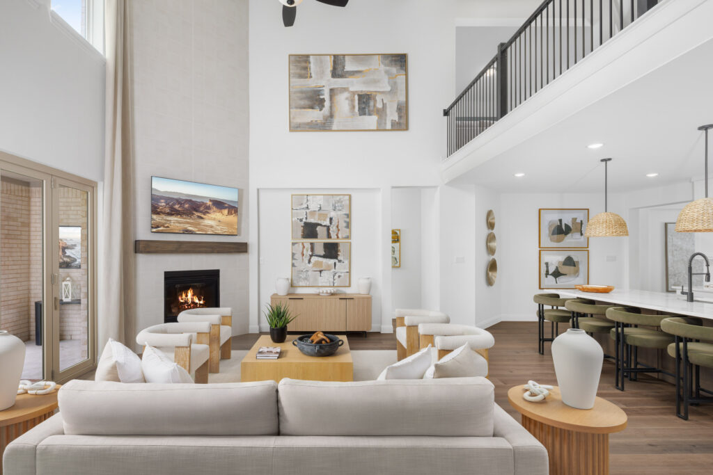 Modern living room with neutral decor, a fireplace, wall art, and an open layout leading to a kitchen with a long island and barstools. Enjoy high ceilings, large windows, and natural light in this Coventry Homes new home at Painted Tree.