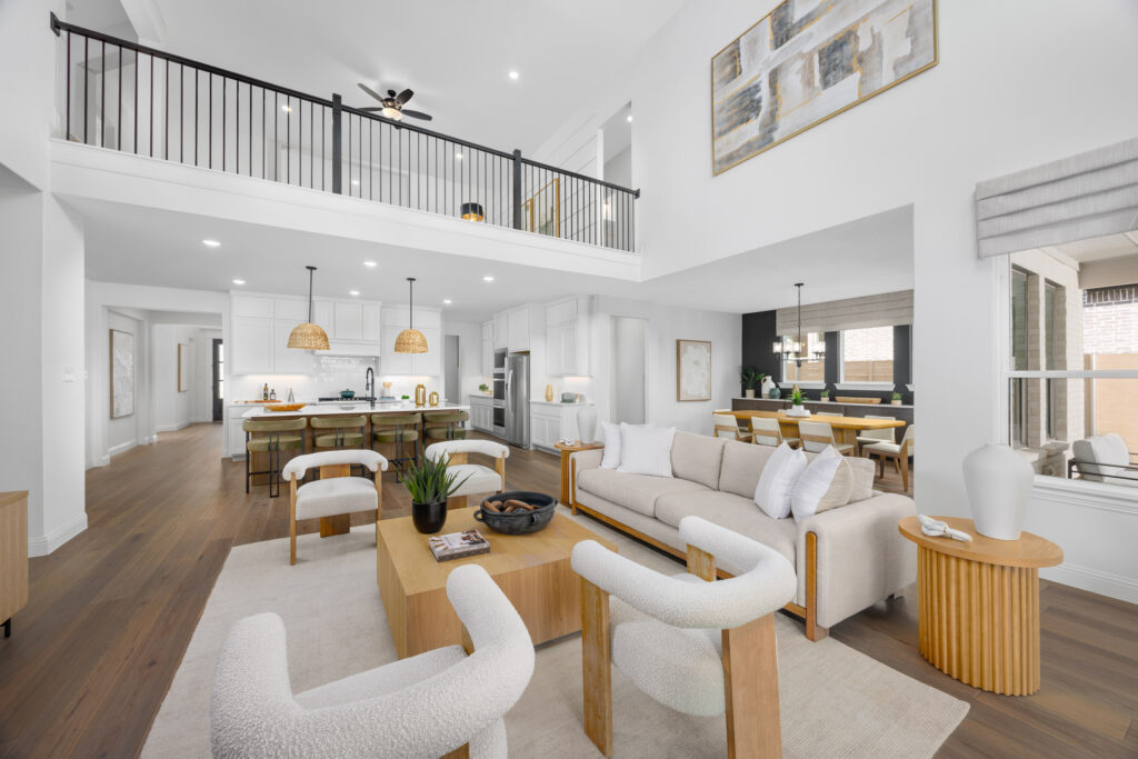 Modern open-concept living area in this new TX home at Painted Tree features white walls, light wood furniture, a stylish kitchen island, dining area, and an upper loft with sleek black railings.