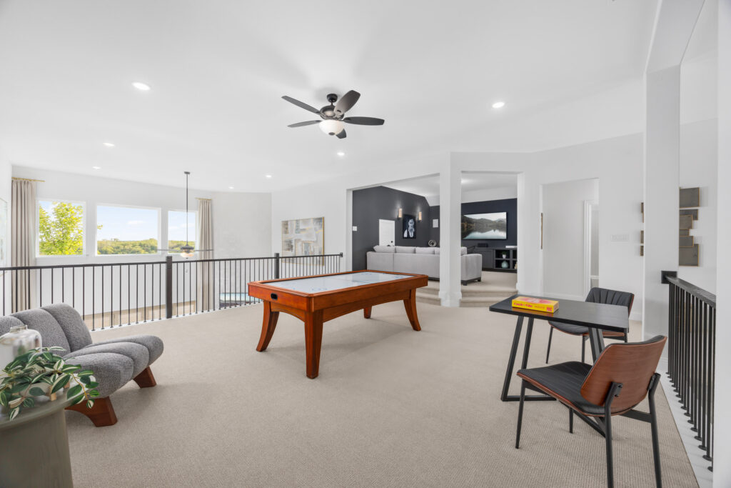 A spacious, modern loft by Coventry Homes in Painted Tree, McKinney features a pool table, two gray chairs, a small desk with two chairs, and an open view into the living area. Large windows provide natural light.