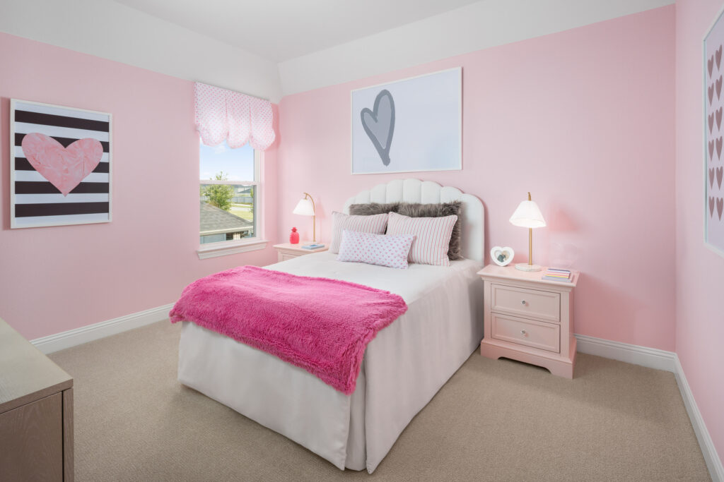 A neatly arranged pink and white bedroom in TX, featuring a bed, bedside tables, lamps, heart-themed wall art, and a window with a patterned curtain—perfect for settling into your new home at Painted Tree.