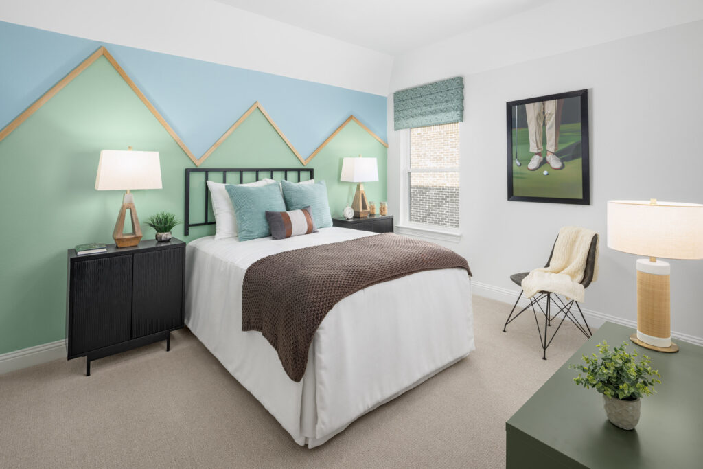 A modern bedroom in McKinney’s Painted Tree community features a double bed, geometric mountain wall art, side tables with lamps, a window, framed golfing picture, and a green bench with lamp and chair—perfect for your new home.