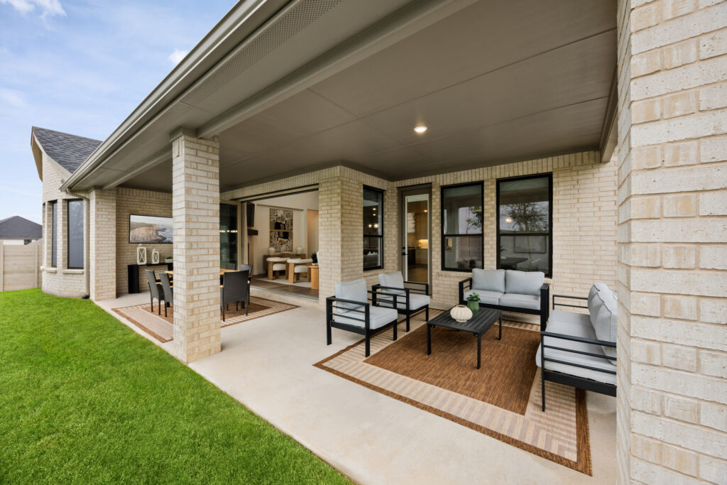 Covered patio attached to a modern brick Coventry Homes house in Painted Tree, McKinney, featuring outdoor seating, rugs, and a view into the dining and living areas through large windows and open doors.