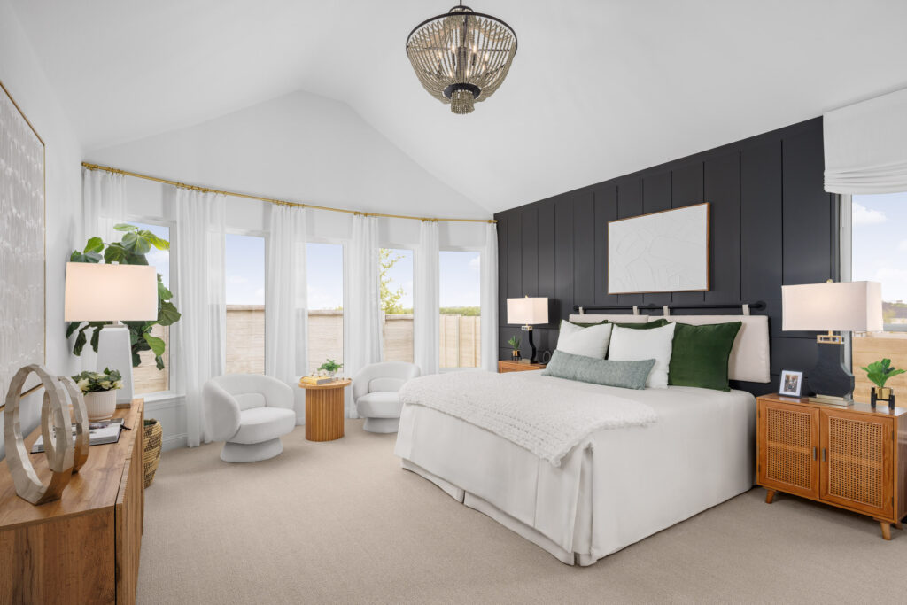 Modern bedroom in a new home in McKinney, TX, featuring a vaulted ceiling, dark accent wall, large bed with white and green bedding, two armchairs by windows, and wooden furniture. Natural light fills the space.