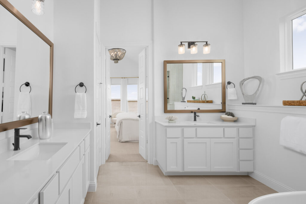 Modern white bathroom with dual vanities, large mirrors, black fixtures, and neutral tile floor in a new home in McKinney, TX. Enjoy a view into a bright, neatly made bedroom through open doors.
