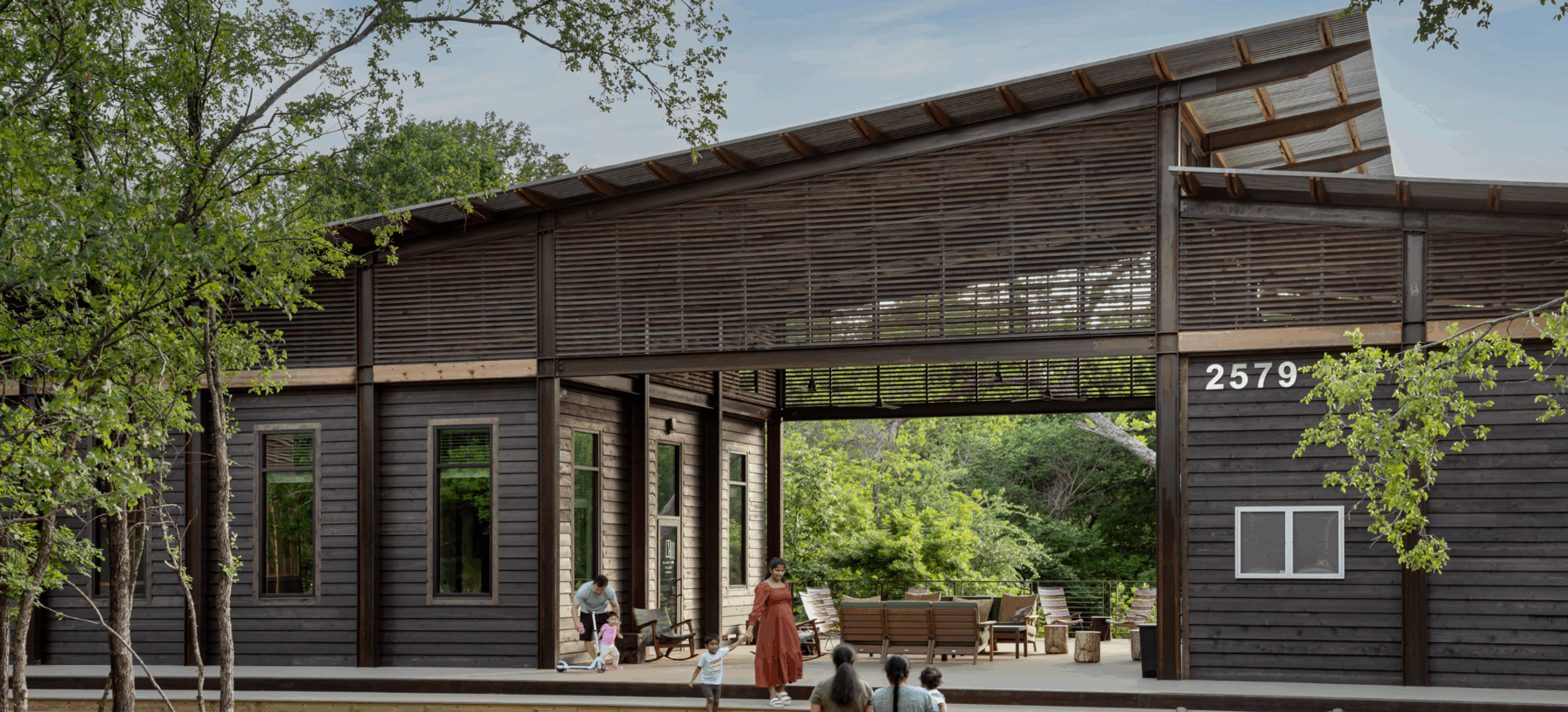 A modern, dark wood pavilion called The Outpost features large open spaces and a slatted roof, nestled among trees in McKinney. Several people stand and sit near the entrance, with building number 2579 visible—an inviting spot near new homes.