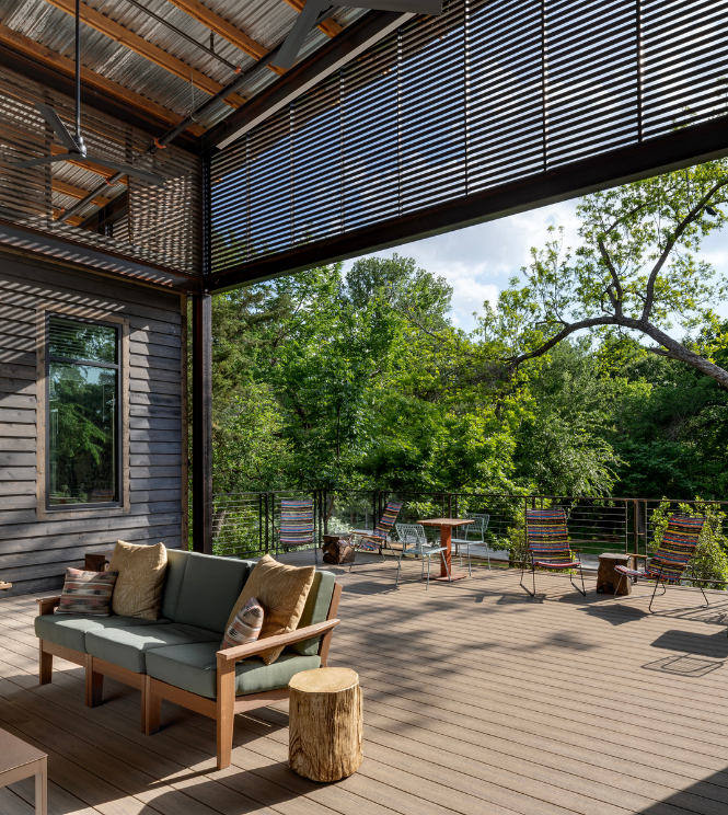 Modern outdoor patio with wooden deck, cushioned sofa, stump side table, and lounge chairs under a partially covered roof—perfect for relaxing in the tranquil setting of Painted Tree, McKinney’s premier community for new homes.