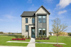 Two-story modern house with gray brick exterior, large windows, and a small front garden, set on a green lawn under a blue sky with a few clouds—one of the stunning new homes by Normandy Homes in McKinney.