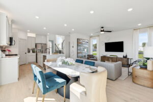 Open-concept living room, dining area, and kitchen by Normandy Homes in Painted Tree, McKinney, with modern furnishings, light wood flooring, and neutral colors; blue and cream chairs surround a marble dining table.