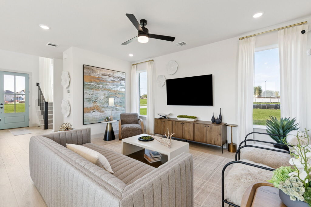 Modern living room in McKinney with neutral furniture, wall-mounted TV, large artwork, and abundant natural light from tall windows; view of entryway and staircase in background—crafted by Normandy Homes at Painted Tree.