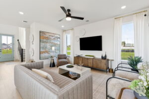 Modern living room in McKinney with neutral furniture, wall-mounted TV, large artwork, and abundant natural light from tall windows; view of entryway and staircase in background—crafted by Normandy Homes at Painted Tree.