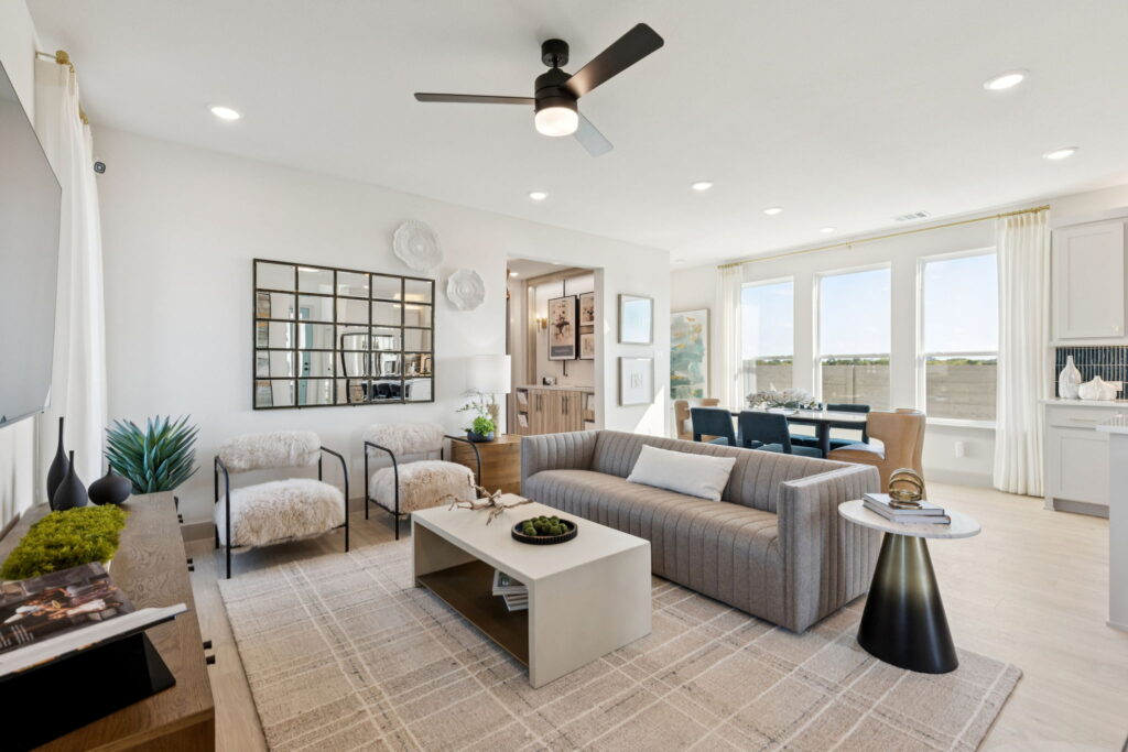 Modern living room with a gray sofa, two armchairs, a coffee table, and a wall mirror, adjacent to a dining area with large windows and neutral decor—showcasing the stylish design of new homes by Normandy Homes in McKinney.
