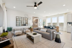 Modern living room with a gray sofa, two armchairs, a coffee table, and a wall mirror, adjacent to a dining area with large windows and neutral decor—showcasing the stylish design of new homes by Normandy Homes in McKinney.