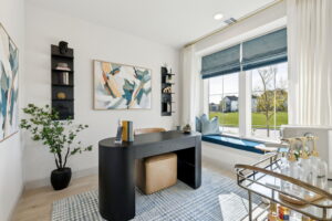 Modern home office in one of the new homes by Normandy Homes at Painted Tree, featuring a black desk, tan chair, abstract wall art, shelves, potted plant, window seat, and a large window overlooking a suburban street.