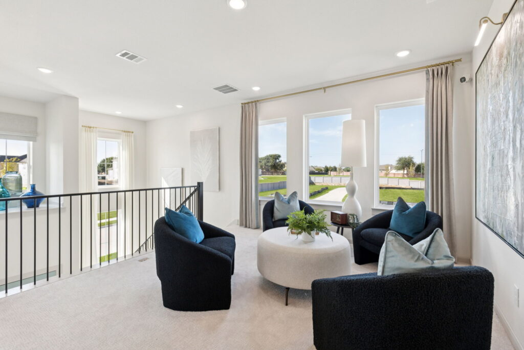 Modern sitting area with three black armchairs, a round white ottoman, large windows with beige curtains, and a white lamp, overlooking a green outdoor space—perfect for relaxing in stylish Normandy Homes at Painted Tree.