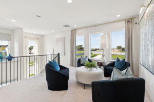 Modern sitting area with three black armchairs, a round white ottoman, large windows with beige curtains, and a white lamp, overlooking a green outdoor space—perfect for relaxing in stylish Normandy Homes at Painted Tree.