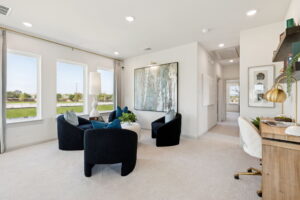 Modern living room in Painted Tree by Normandy Homes, featuring three black chairs around a white table, large artwork on the wall, a desk with lamp, and several windows filling the space in these new homes with natural light.
