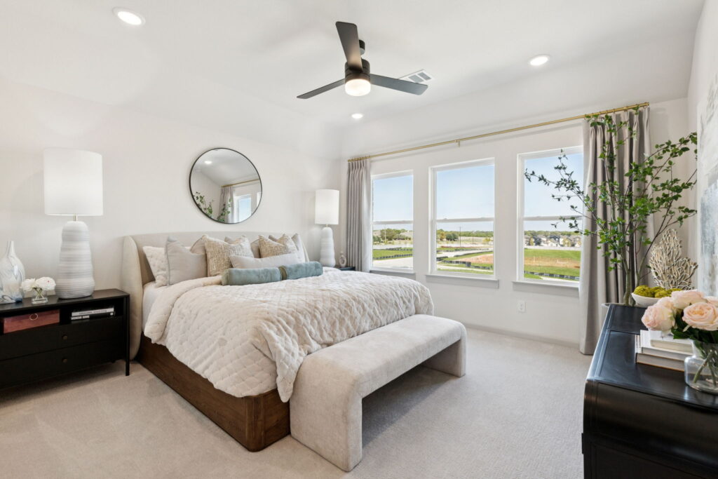 A modern, bright bedroom in one of the stunning new homes by Normandy Homes in McKinney, featuring a large bed, bench, nightstands, ceiling fan, and windows with scenic outdoor views.