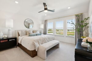 A modern, bright bedroom in one of the stunning new homes by Normandy Homes in McKinney, featuring a large bed, bench, nightstands, ceiling fan, and windows with scenic outdoor views.
