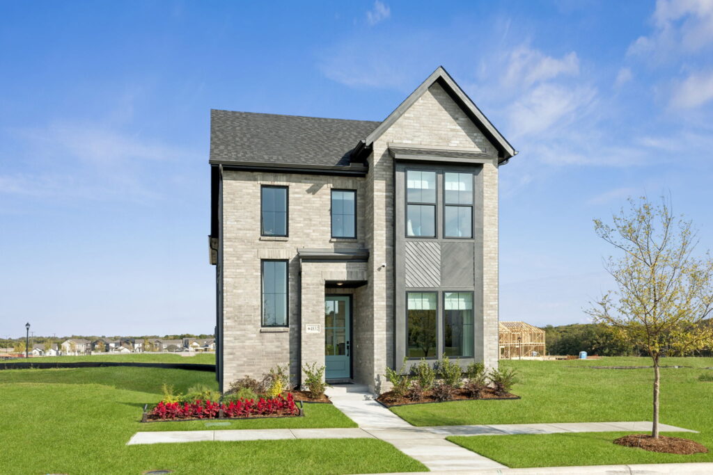 Two-story modern house with gray brick exterior, large windows, and manicured lawn in McKinney’s Painted Tree community. Enjoy new homes with small garden beds, set in a suburban neighborhood under a clear blue sky.