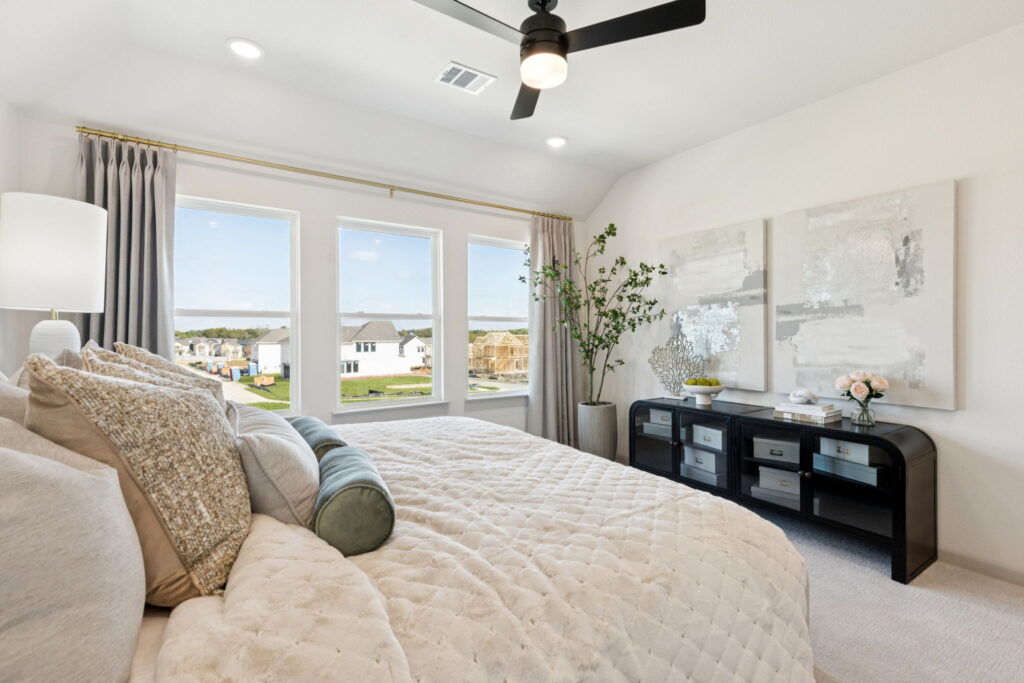 A modern bedroom in Normandy Homes features a large bed, neutral bedding, three windows, a ceiling fan, a plant, abstract wall art, and a black dresser with decor—perfect for your new home in Painted Tree.