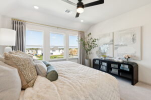 A modern bedroom in Normandy Homes features a large bed, neutral bedding, three windows, a ceiling fan, a plant, abstract wall art, and a black dresser with decor—perfect for your new home in Painted Tree.