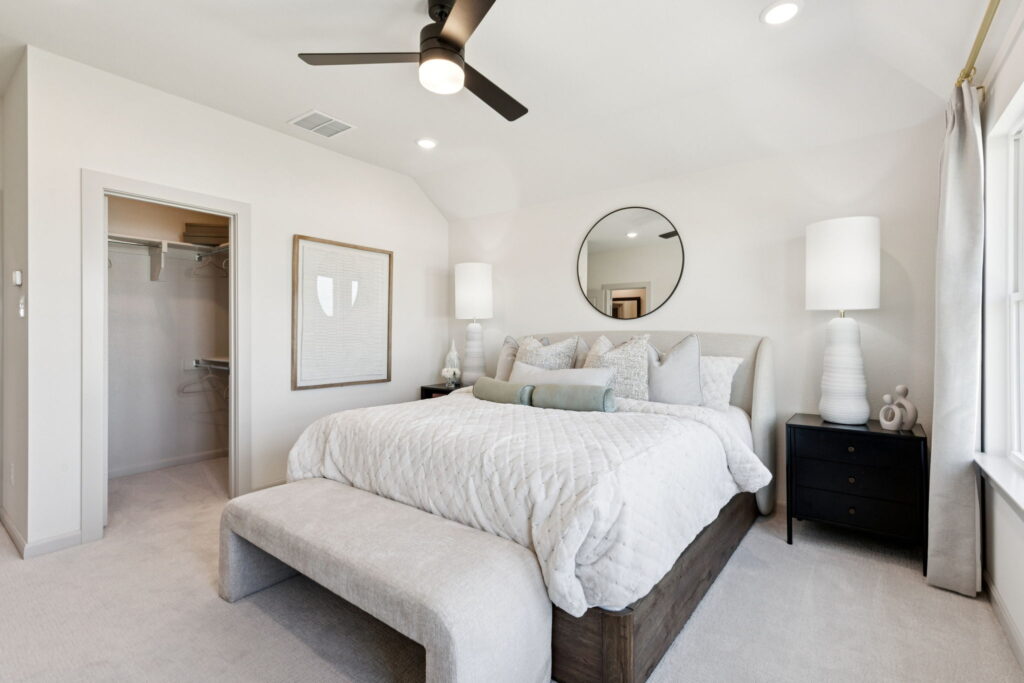A modern bedroom in McKinney by Normandy Homes features a large bed, black nightstands, white lamps, a round mirror, ceiling fan, and open walk-in closet. Light walls and natural light from a window complete this new home retreat.