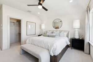 A modern bedroom in McKinney by Normandy Homes features a large bed, black nightstands, white lamps, a round mirror, ceiling fan, and open walk-in closet. Light walls and natural light from a window complete this new home retreat.