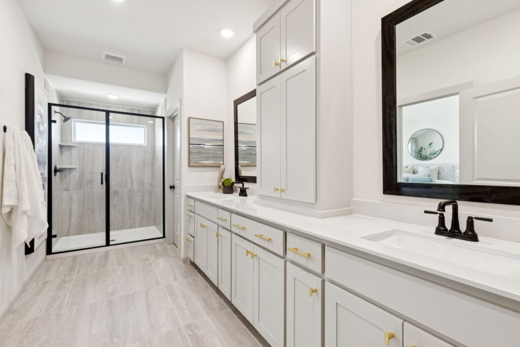 Modern bathroom in Painted Tree, McKinney, with double sinks, white cabinets with gold hardware, a glass-enclosed shower, light tile flooring, and large wall mirrors by Normandy Homes.