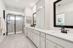 Modern bathroom in Painted Tree, McKinney, with double sinks, white cabinets with gold hardware, a glass-enclosed shower, light tile flooring, and large wall mirrors by Normandy Homes.
