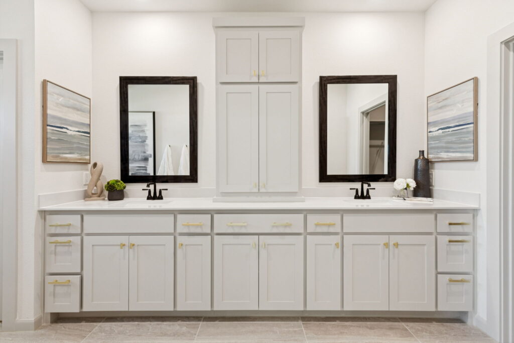 Double-sink vanity with white cabinets, two mirrors, brass hardware, and decorative items; neutral-toned artwork on walls and tiled floor add style to this Painted Tree new home in McKinney.