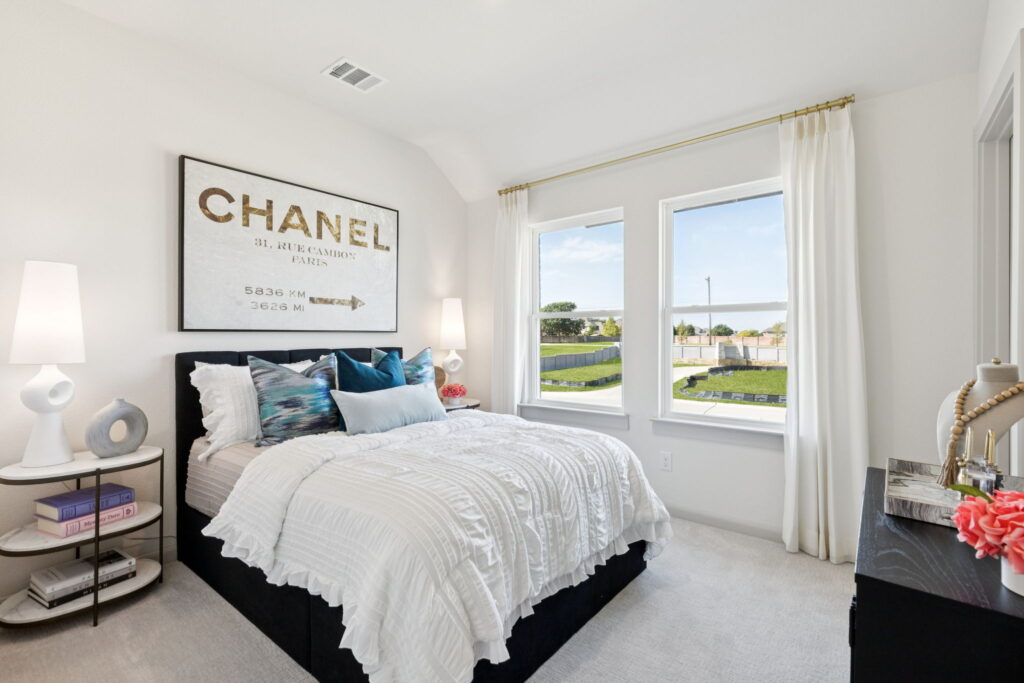 A modern bedroom in a Normandy Homes residence features a neatly made bed, decorative pillows, a Chanel art print above the headboard, two windows with white curtains, and a black dresser with decor items—perfect for stylish living in McKinney.