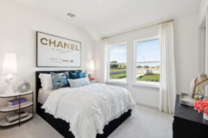 A modern bedroom in a Normandy Homes residence features a neatly made bed, decorative pillows, a Chanel art print above the headboard, two windows with white curtains, and a black dresser with decor items—perfect for stylish living in McKinney.