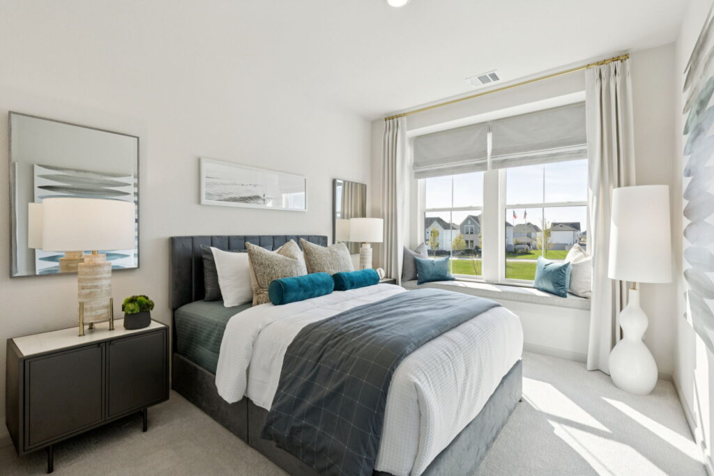 A modern bedroom by Normandy Homes in Painted Tree, McKinney, features a made bed, two bedside tables with lamps, a large window with a window seat, and light-colored walls and curtains.