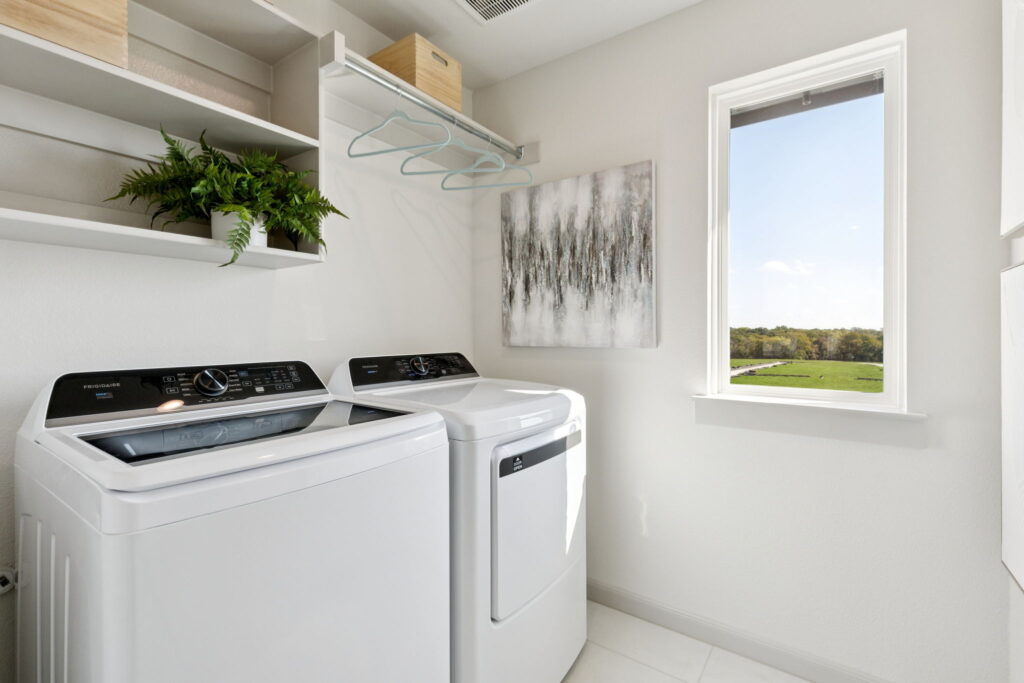 A laundry room in Painted Tree, McKinney, features a washing machine, dryer, wall shelves, hanging rods with hangers, a potted plant, abstract wall art, and a window showcasing the outdoor view—perfect for new homes.
