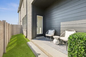 A cozy outdoor patio with two chairs and a side table on a concrete slab, adjacent to a Normandy Homes residence in McKinney, featuring dark siding and enclosed by a wooden privacy fence.