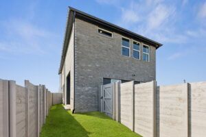Two-story modern brick house with three windows on the upper floor, located in Painted Tree, McKinney. Surrounded by a tall white fence and green lawn under a clear blue sky, it's perfect for those seeking new homes in a vibrant community.